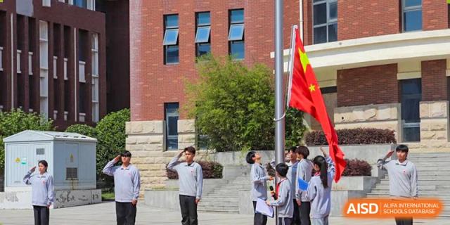 Students standing near a flagpole with the Chinese flag and school buildings in the background during a ceremony at Glocal Elite International School.