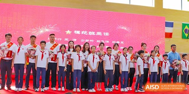 A group of students smiling and holding trophies or flowers on a stage with a bright red backdrop during an awards ceremony at Glocal Elite International School.