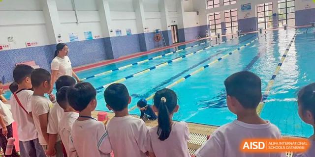 Students from behind, wearing white shirts, looking down at the water of the indoor swimming pool during a lesson or practice at Glocal Elite International School.