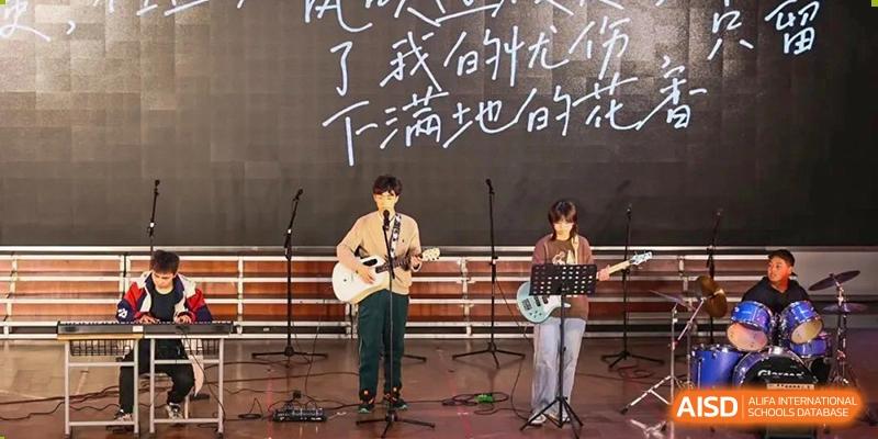 Two students playing acoustic guitars and singing on a stage with Chinese characters written on a large chalkboard backdrop at Maple Leaf International School-Zhenjiang.