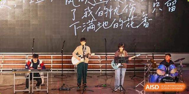 Two students playing acoustic guitars and singing on a stage with Chinese characters written on a large chalkboard backdrop at Maple Leaf International School-Zhenjiang.