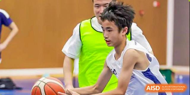 A coach wearing a yellow vest guides a male student on the basketball court during practice at Harrow LiDe School Hengqin.
