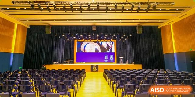Interior wide shot of the Harrow LiDe School Hengqin auditorium or hall with rows of yellow chairs and a large screen on the stage.