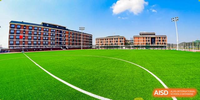 Outdoor view of the Harrow LiDe School Hengqin sports field with bright green turf and traditional brick school buildings in the background.