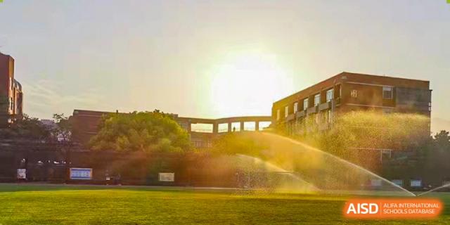 An outdoor sports field being watered with sprinklers, with a building in the background at sunset at the High School Affiliated to Shanghai Jiao Tong University.