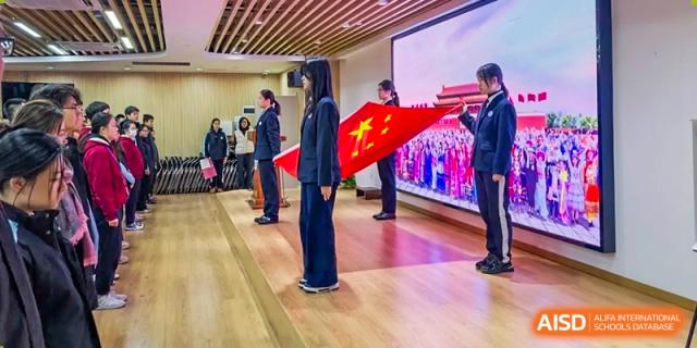 Students in uniform holding the Chinese flag during a ceremony on an indoor stage at the High School Affiliated to Shanghai Jiao Tong University.