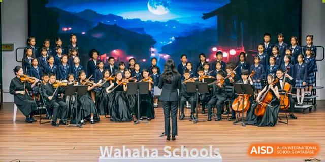 The orchestra from Wahaha Bilingual School Hangzhou is performing on a stage with a scenic backdrop of mountains and a full moon.