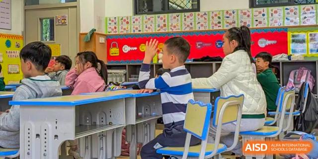 A young male student from Beanstalk International Bilingual School Kunming wearing a striped shirt and enthusiastically raising his hand in a bright classroom.