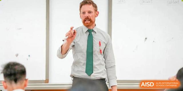 A male teacher at Beanstalk International Bilingual School Kunming wearing a white shirt and green tie, standing in front of a whiteboard and holding a marker.