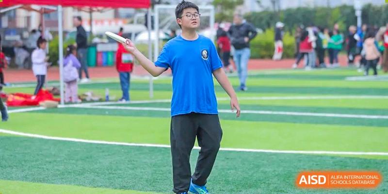 A male student from Beanstalk International Bilingual School Kunming wearing a blue shirt and black pants, throwing a disc or frisbee on a sports field.
