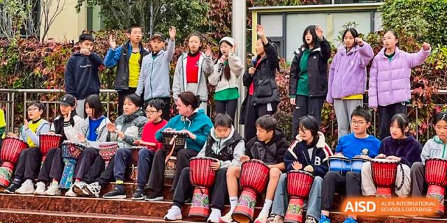 A group of students from Beanstalk International Bilingual School Kunming sitting on stairs. Some are playing djembe drums while others stand and cheer.