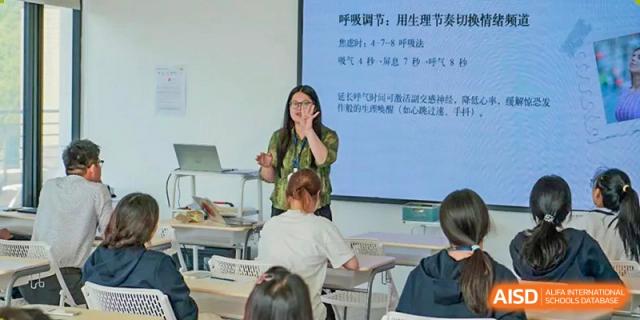 A female teacher from Adcote School Shanghai is giving a presentation in a classroom, pointing to a whiteboard with Chinese characters.