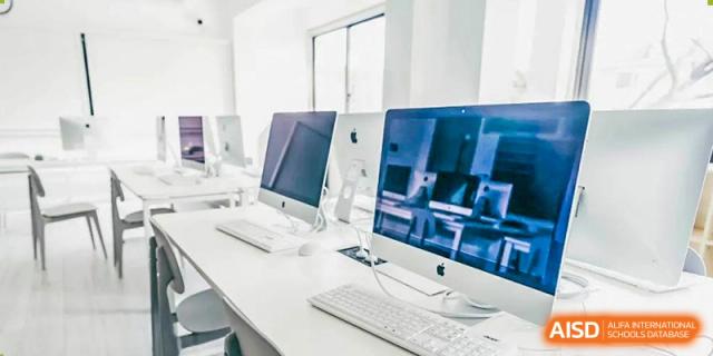 The computer lab at Adcote School Shanghai, with several white computers on desks, is shown from a wide shot.