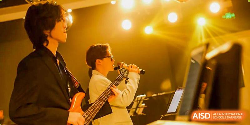 A male student from Adcote School Shanghai sings and plays a bass guitar under bright stage lights during a performance.
