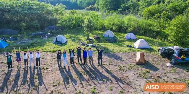 A group of students from Adcote School Shanghai stands outside in a field with tents in the background, celebrating an outdoor camp or event.
