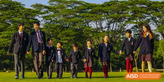 A group of diverse Bromsgrove School Mission Hills students in uniform holding hands and running on a grassy field with trees in the background.