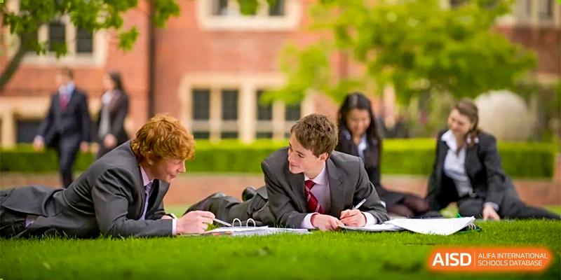 Two students at Bromsgrove School Mission Hills are sitting outside on the grass, taking notes with a laptop and books, with other students blurred in the background.
