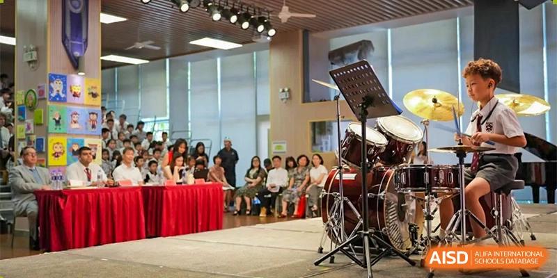An audience watching a musical performance on a stage, with a drum set visible, at Bromsgrove School Mission Hills.