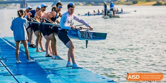 A student in white and blue uniform carefully carrying a rowing shell to the water from a blue pontoon, representing the rowing program at Harrow Lide High School Nanning.