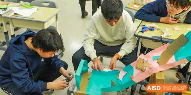 A close-up of a student in a white sweater working on an engineering or design project using green and blue materials at Guokai International School of Qingdao.