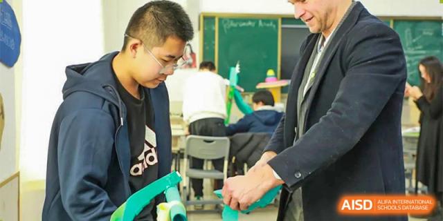 Students and an adult are working together on a hands-on project or experiment, with a green chalkboard visible in the background, at Guokai International School of Qingdao.