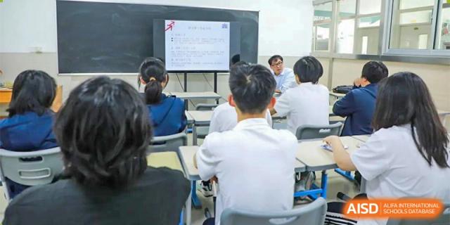 Students are seated, viewing a presentation or lesson displayed on a large screen at the front of the classroom at Guokai International School of Qingdao.