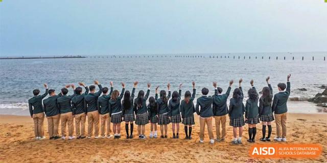 A large group of students in school uniform standing on a beach with their hands raised against the backdrop of the sea and sky at Guokai International School of Qingdao.