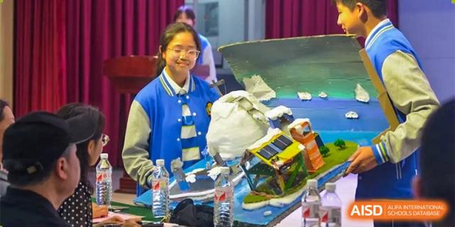 A young woman in a blue and white jacket is smiling next to a science project at Chengdu Meishi International School.