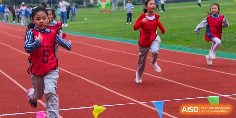 A young girl in a red hoodie and gray pants runs on a red track with a yellow flag nearby at an event for Chengdu Meishi International School.