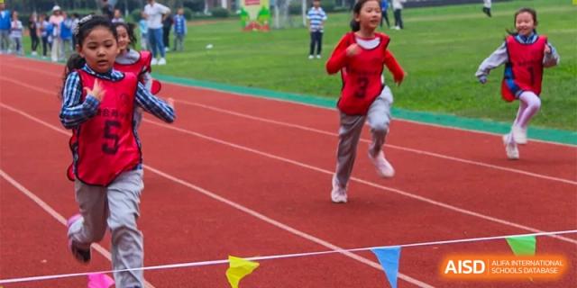A young girl in a red hoodie and gray pants runs on a red track with a yellow flag nearby at an event for Chengdu Meishi International School.
