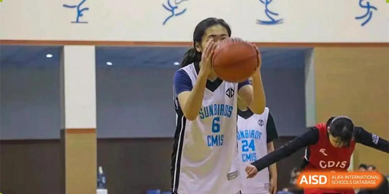 A female student in a white and blue basketball jersey with the number '6' on it is holding a basketball during a game at Chengdu Meishi International School.