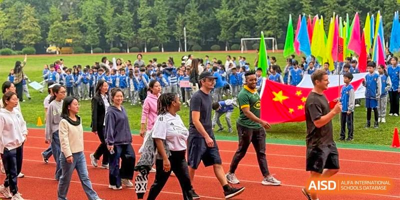 A group of people, including students and adults, are walking in a procession on a sports field, with one person holding a national flag at Chengdu Meishi International School.