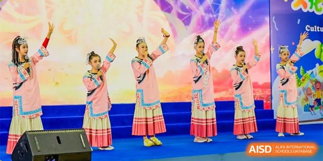 Two female students in traditional, colorful dresses are performing on a stage with their hands raised at Chengdu Meishi International School.