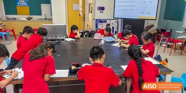Students from Tsinglan School sitting at a large table and working on a project with laptops and tablets.