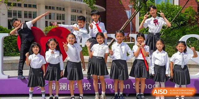 A group of young students from Tsinglan School, in their uniforms, posing with teachers in front of a school banner.