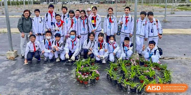 A group of students from Shanghai Foreign Language School Affiliated to SISU wearing red scarves and gloves, engaging in a gardening or planting activity outdoors.