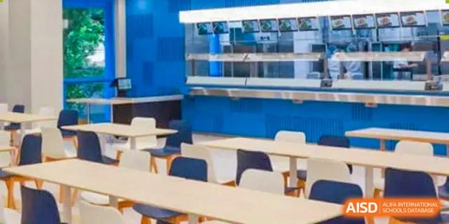 A modern, well-lit cafeteria or dining area with light wood tables and a blue service counter at Shanghai Foreign Language School Affiliated to SISU.