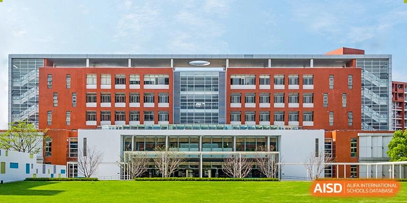 The main exterior building of Vanke School Pudong, a modern, multi-story structure with red brick accents and large windows, fronted by a green lawn and clear blue sky.