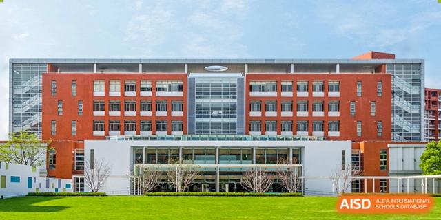 The main exterior building of Vanke School Pudong, a modern, multi-story structure with red brick accents and large windows, fronted by a green lawn and clear blue sky.