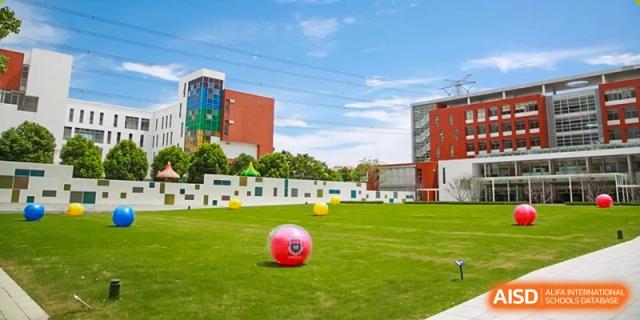 An outdoor green playing field at Vanke School Pudong with a large red ball, set against the modern school buildings and a blue sky.