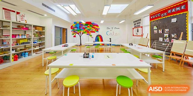 A bright art or classroom space at Vanke School Pudong with a long white table, small green stools, and a colorful tree mural on the wall.