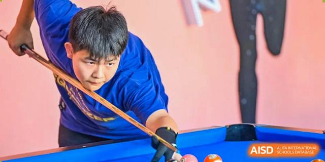 A male student wearing a blue shirt and a black glove is focused on a pool game, lining up a shot on a blue felt pool table Suzhou North America High School.