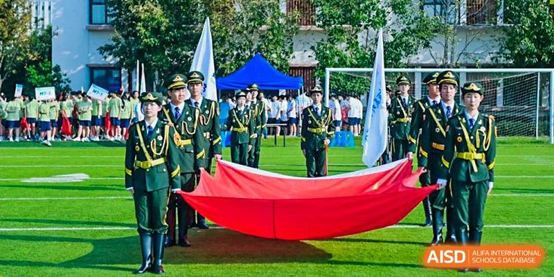 Students from Suzhou North America High School are holding a large red banner during an outdoor event with military personnel in the background.