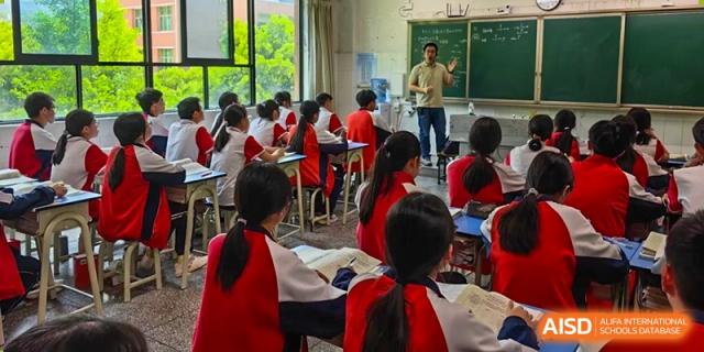 A male teacher from Tianjin Nankai High School  is standing at the front of a classroom, giving a lecture to a group of students in red uniforms.