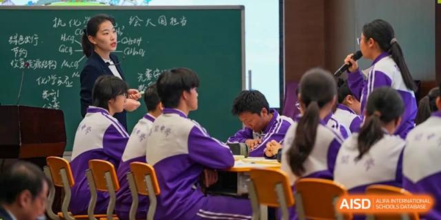 Two students from Tianjin Nankai High School International Department, wearing purple and white uniforms, are seated at a desk and studying together in a classroom.