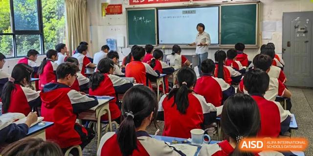 A female teacher from Tianjin Nankai High School  is at the front of a classroom, writing on a whiteboard.