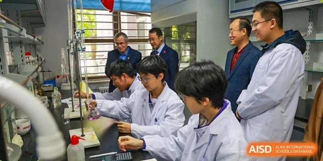 Two male students from Tianjin Nankai High School  are wearing lab coats and seated at a table with a teacher, looking at a laptop.