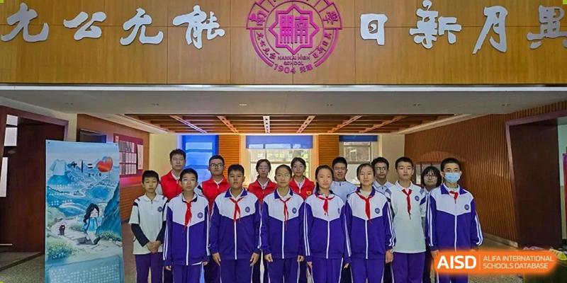 A photo of a group of students from Tianjin Nankai High School  standing together for a photo outside a school building with the school's emblem on the wall behind them.