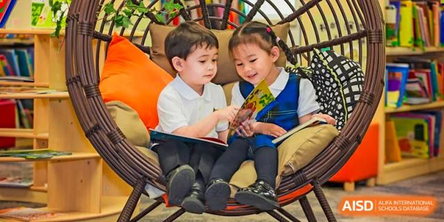 Two young Wellington College International Tianjin students, a boy and a girl in school uniforms, are seated together in a chair and looking at a book.