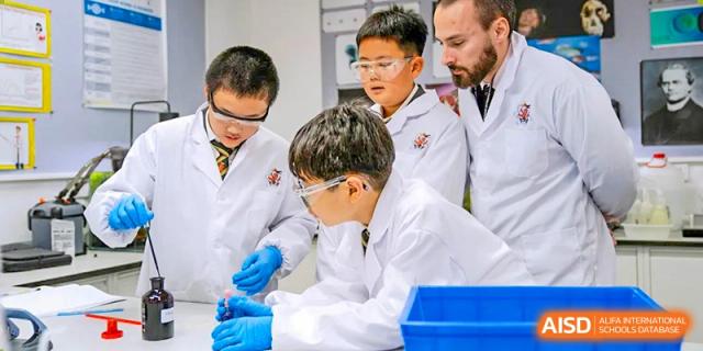 A male teacher from Wellington College International Tianjin supervises two male students in lab coats and safety goggles during an experiment.